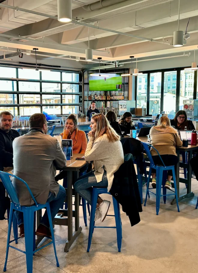Busy indoor bar and dining area with people seated at high-top tables and sports on wall-mounted TVs