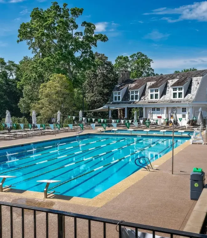 Outdoor lap pool with diving boards, lane lines, and lounge chairs in front of a clubhouse at a private golf club